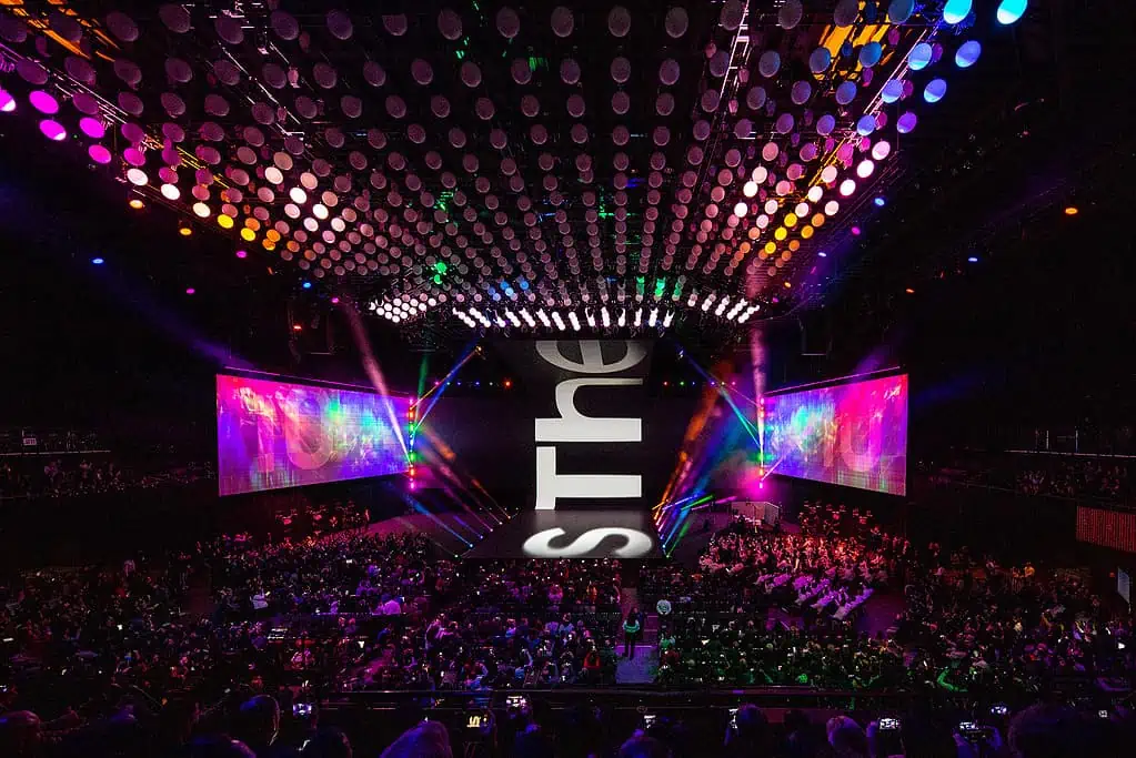 An enthusiastic audience cheers as the band plays their vibrant music on the illuminated stage in the purple, magenta, and violet lit arena.