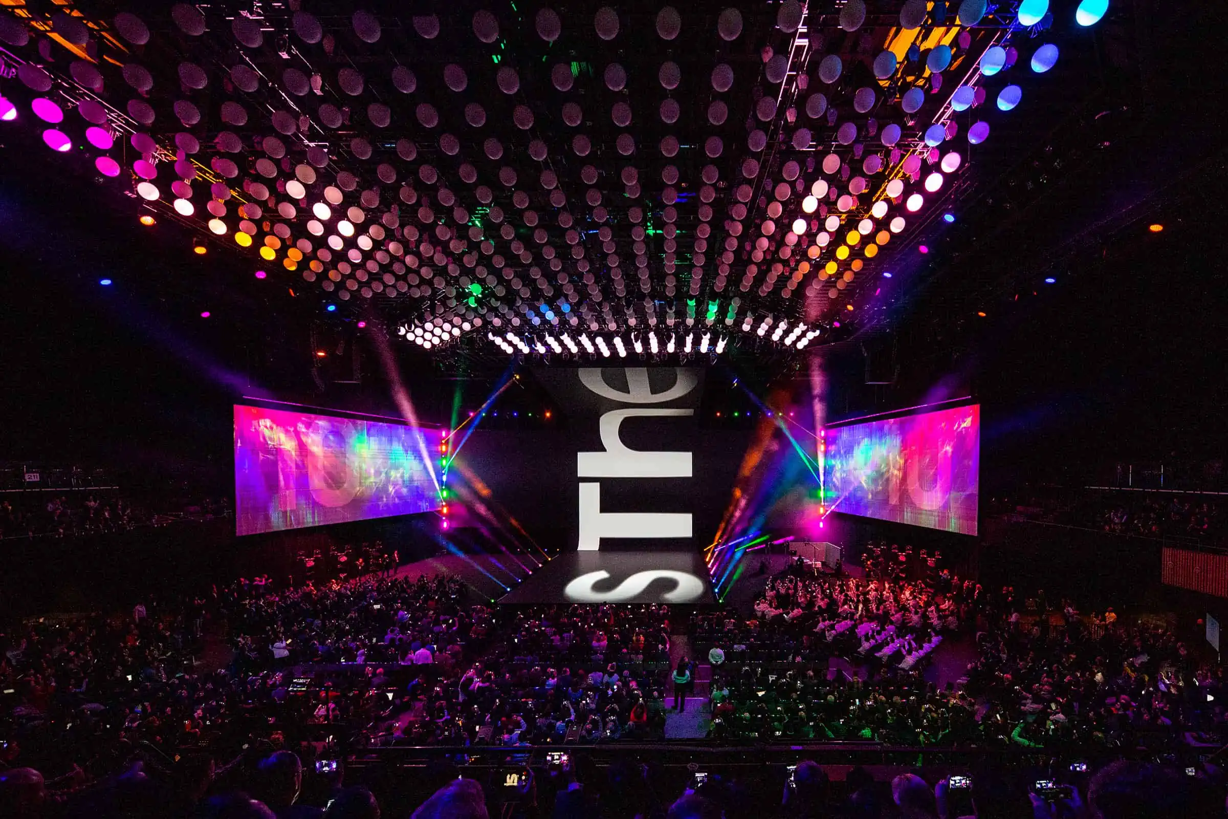 An enthusiastic audience cheers as the band plays their vibrant music on the illuminated stage in the purple, magenta, and violet lit arena.